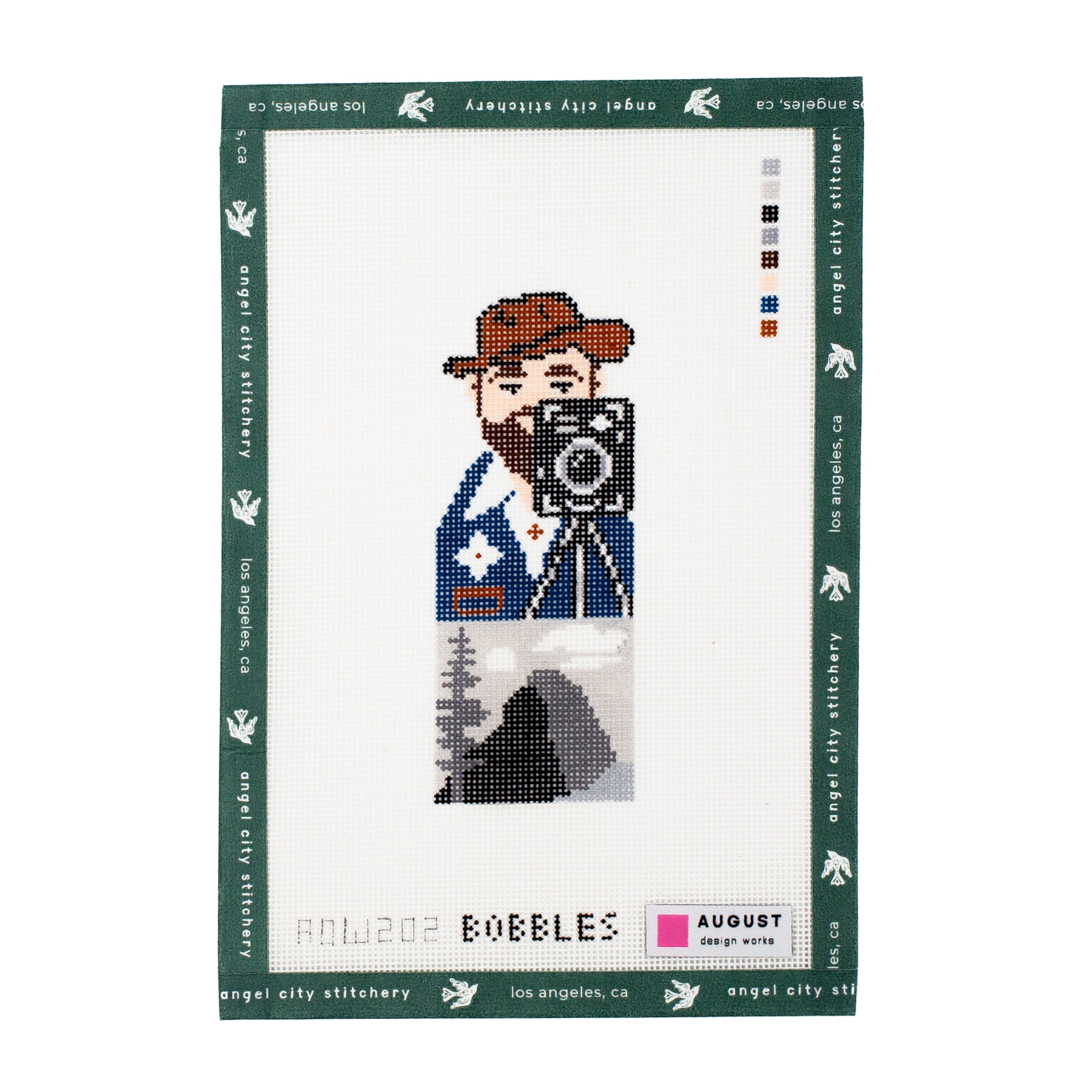Needlepoint canvas pattern packaging with a design of a person holding a camera on a white background.