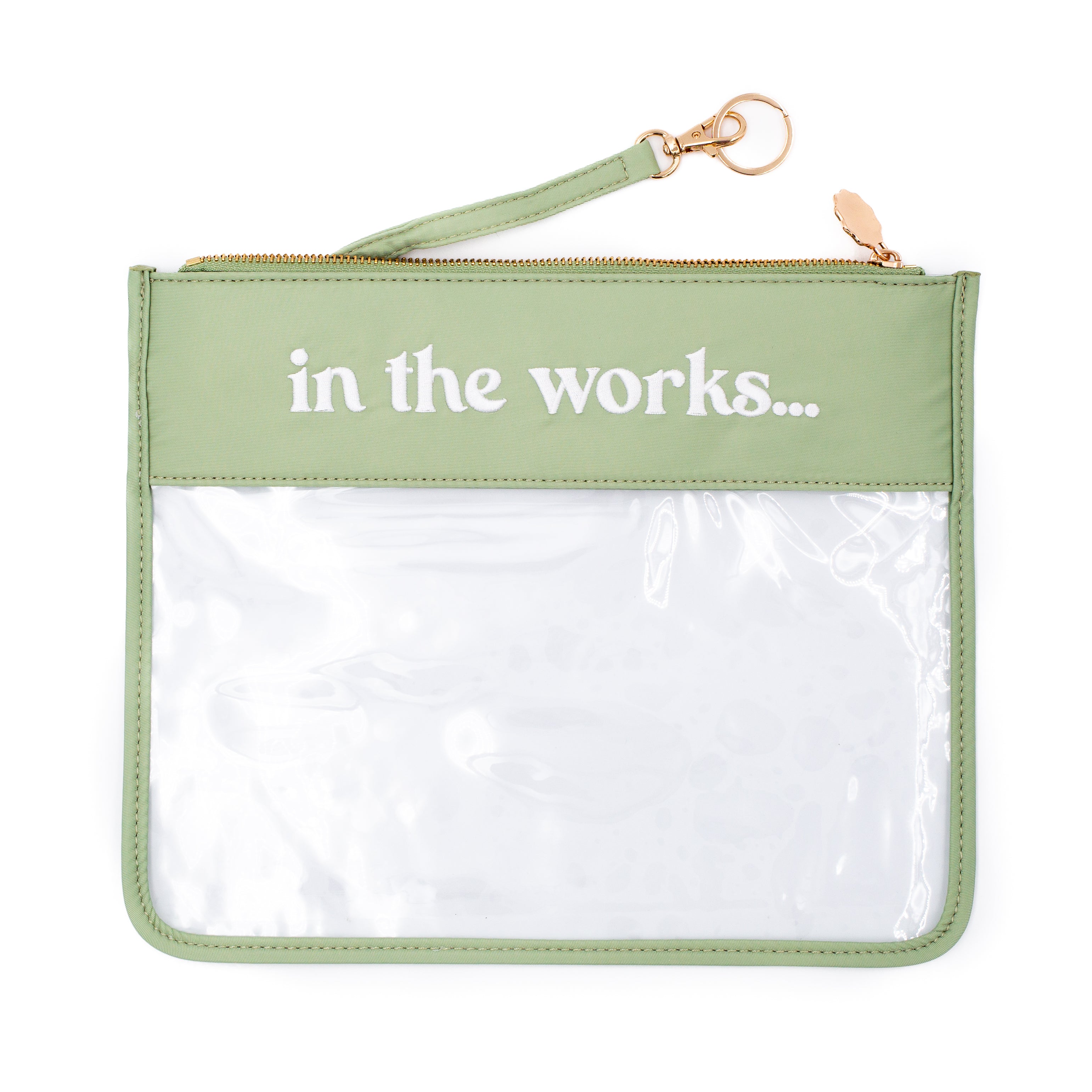A sage green zippered pouch with a clear plastic front window and the words 'in the works.