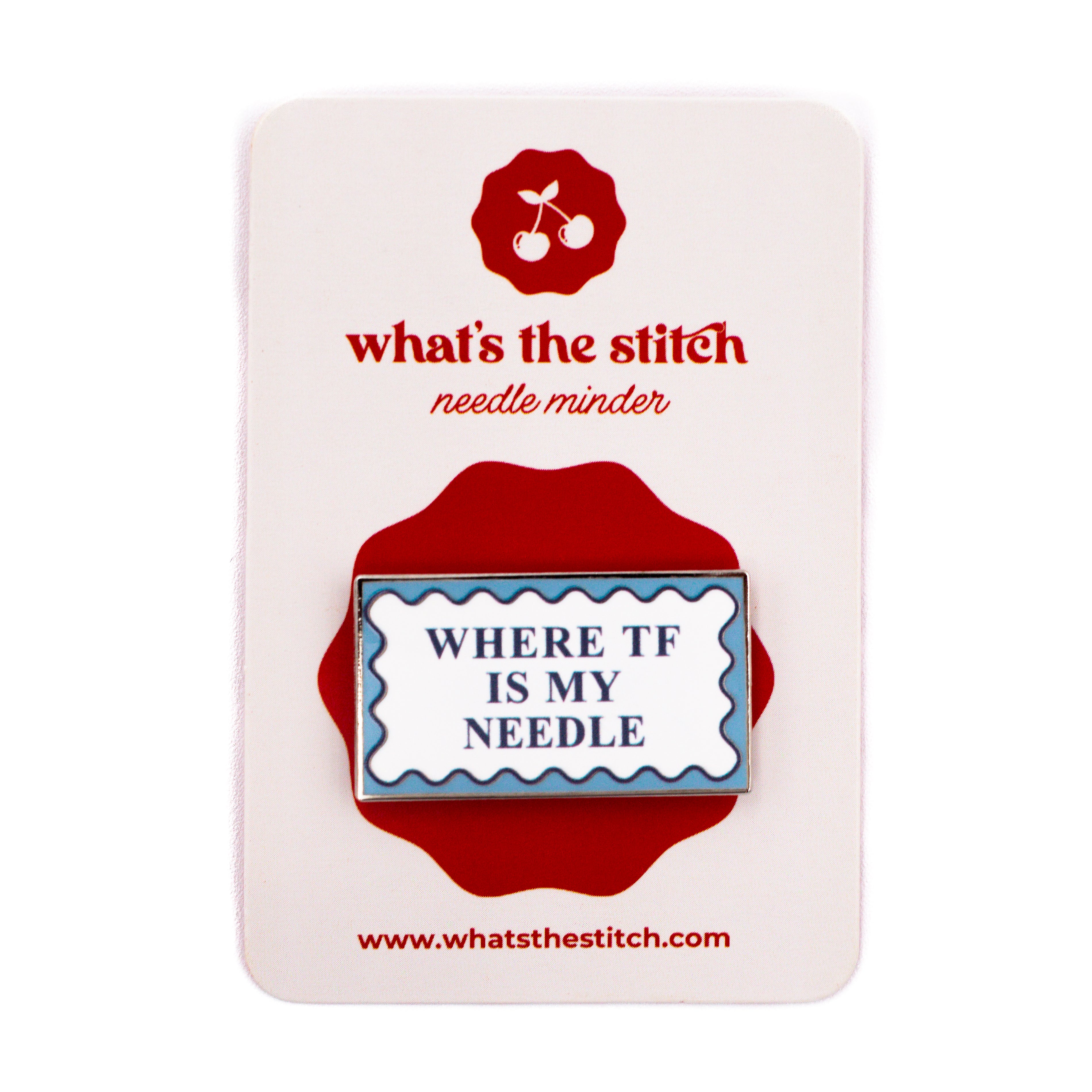 A rectangular enamel needle minder by what's the stitch, featuring the text "WHERE TF IS MY NEEDLE" in dark blue inside a white and blue scalloped border, mounted on a white display card with a red logo and accents