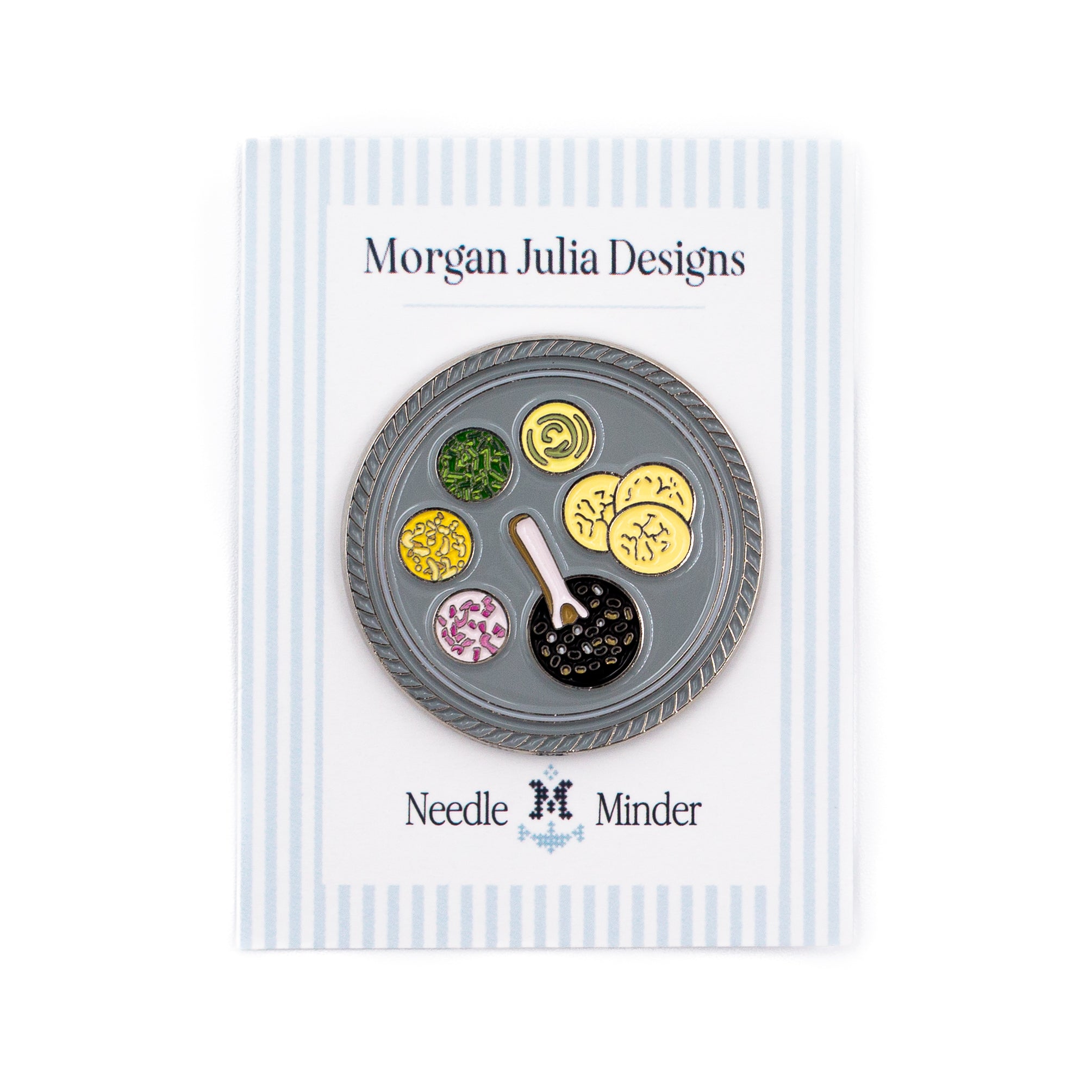 A round enamel needle minder by Morgan Julia Designs, featuring an illustration of a gray tray with various food dishes and a spoon, mounted on a white and blue striped display card