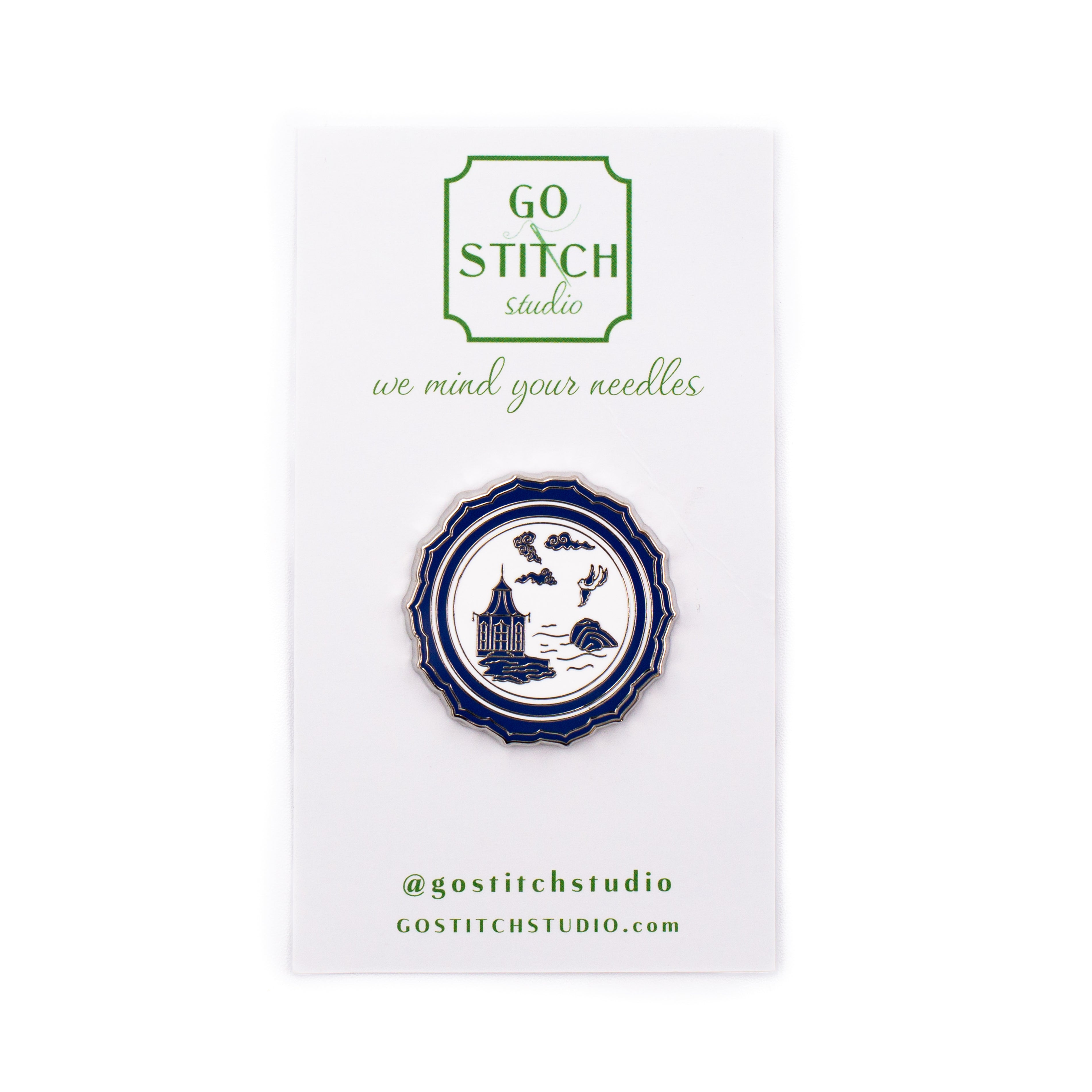 A circular enamel pin by Go Stitch Studio, featuring a navy blue and white illustration of a castle on a ship's wheel, mounted on its original white display card.