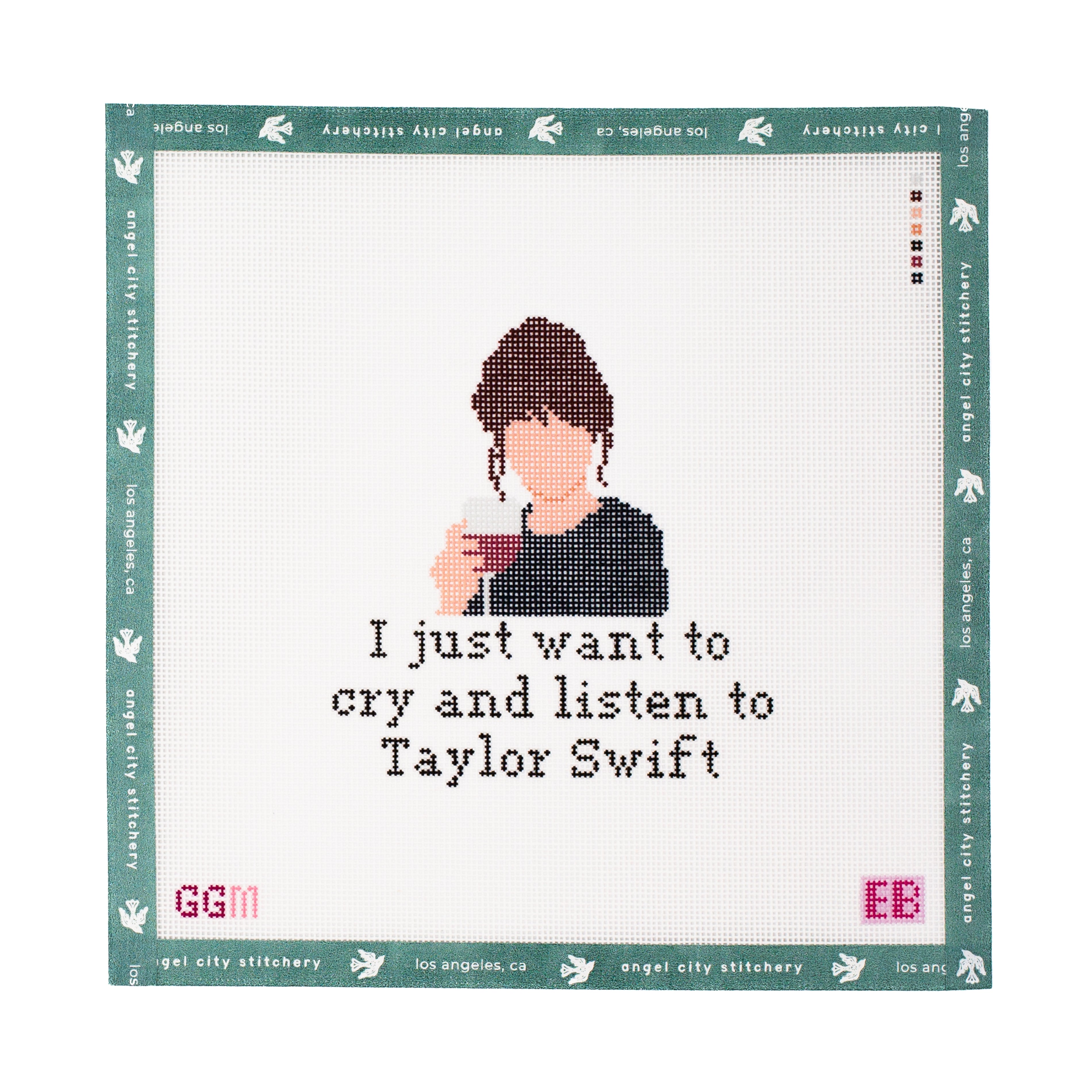 A square canvas cross-stitch design with the text 'I just want to cry and listen to Taylor Swift' stitched below. The design is framed by a teal border with white bird motifs and the text 'Angel City Stitchery', with 'GGM' and 'EB' stitched in pink at the bottom corners.