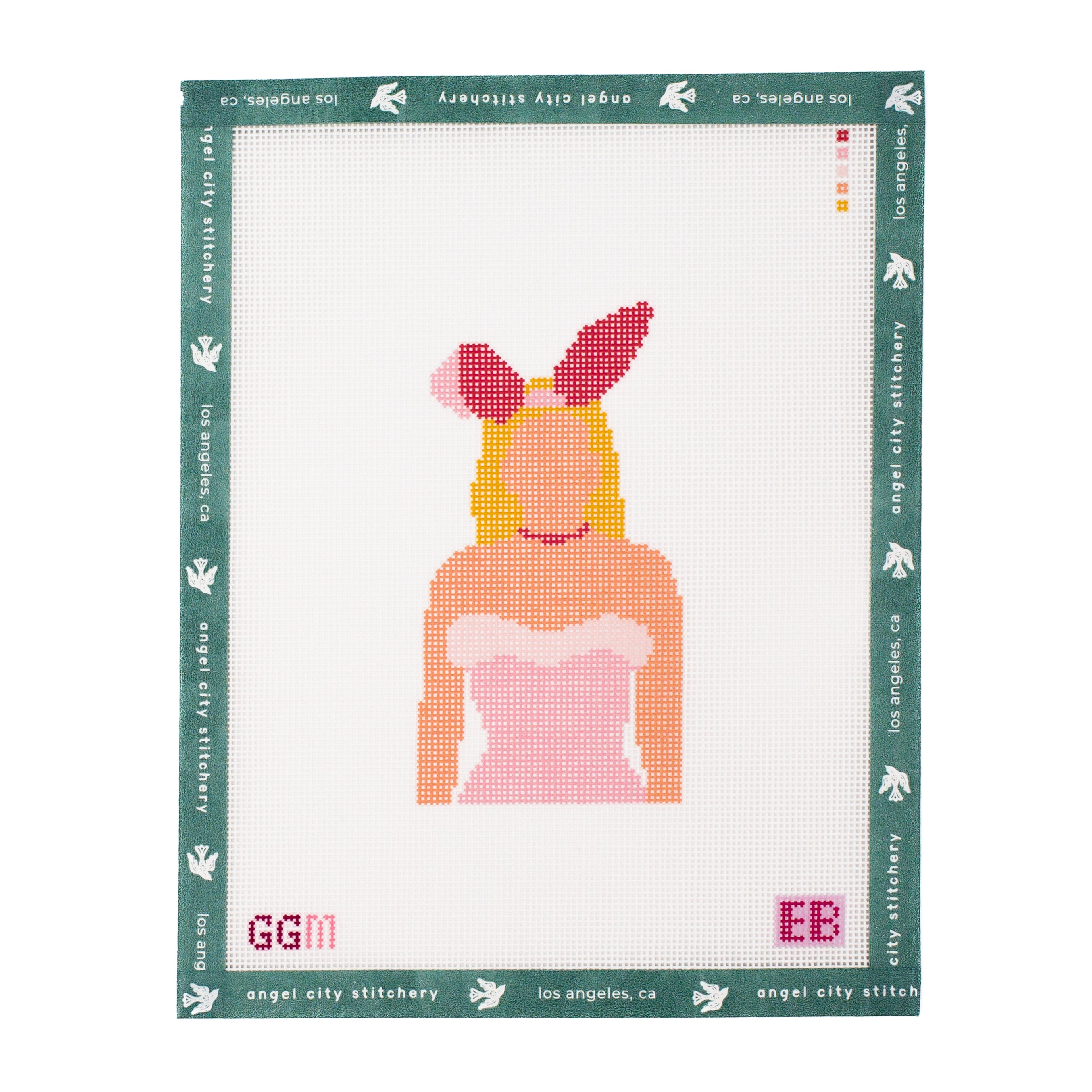 A completed cross-stitch or needlework sampler on white Aida cloth, featuring a stylized bunny girl figure with pink bunny ears, yellow hair, and a pink dress, surrounded by a green border printed with 'angel city stitchery' and 'Los Angeles, ca'.