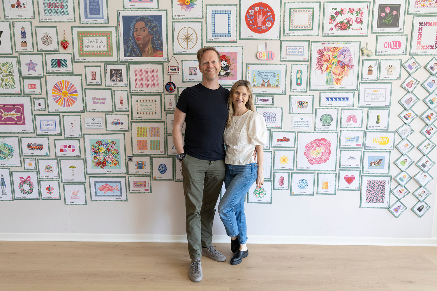 Two people standing in front of a wall filled with various framed needlepoint canvases.