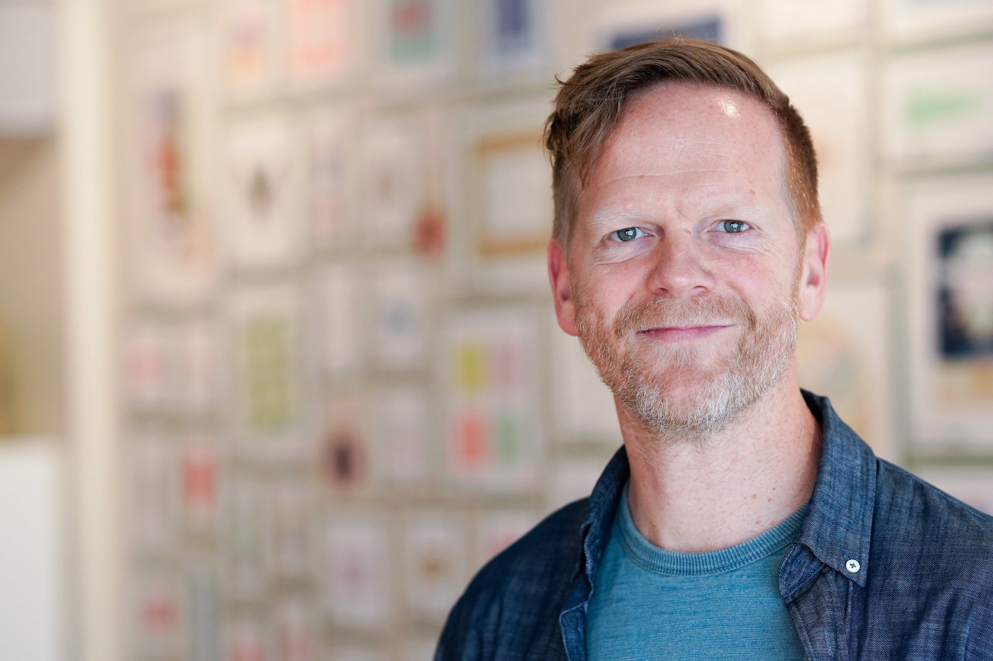 Man with a beard and short hair wearing a blue shirt and dark jacket in front of a colorful wall.