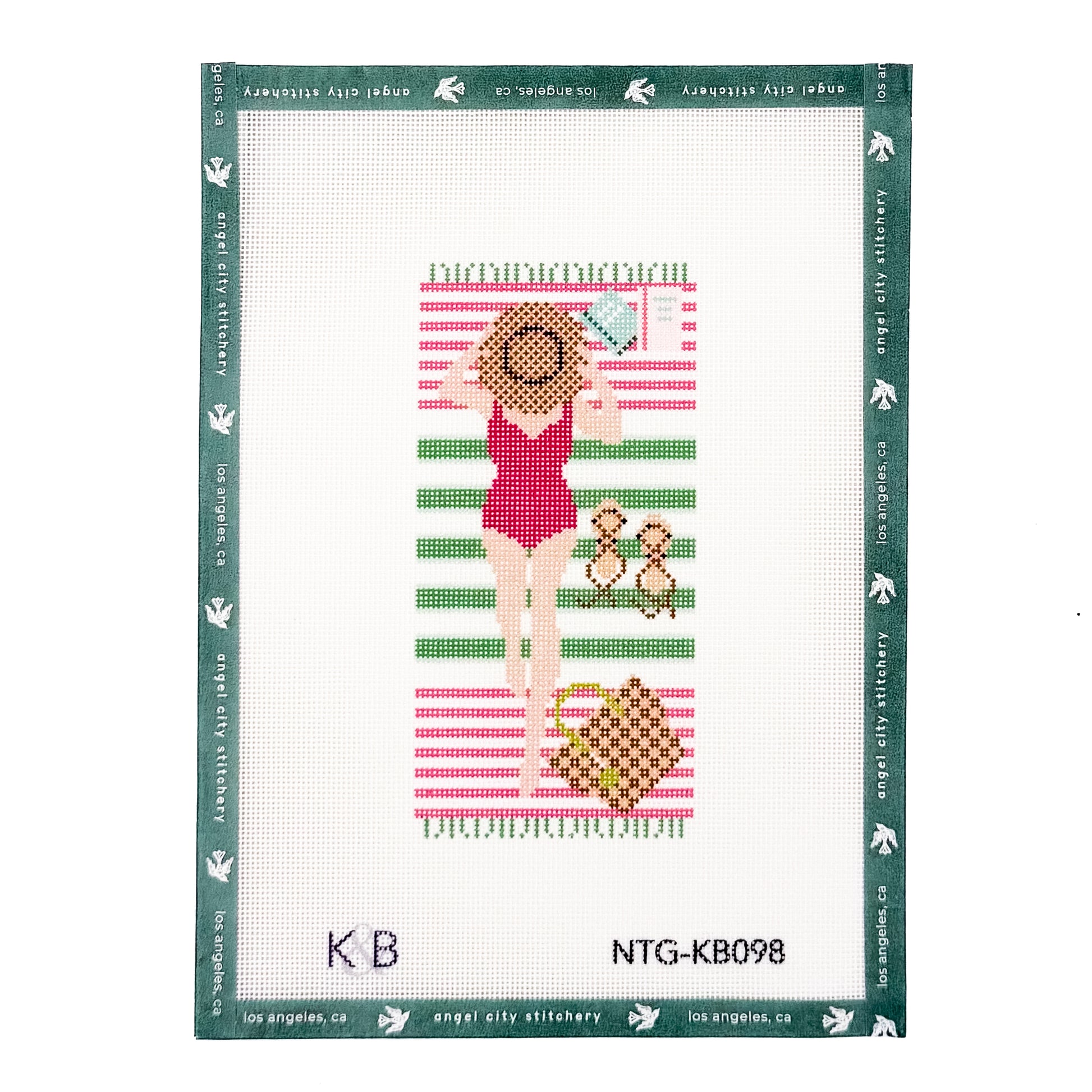 A finished needlepoint canvas featuring a woman in a red swimsuit lying on a pink and green striped beach towel with a hat, sunglasses, and a basket, bordered by a green brand frame. The text 'NTG-KB098' and 'angel city stitchery' are visible at the bottom.