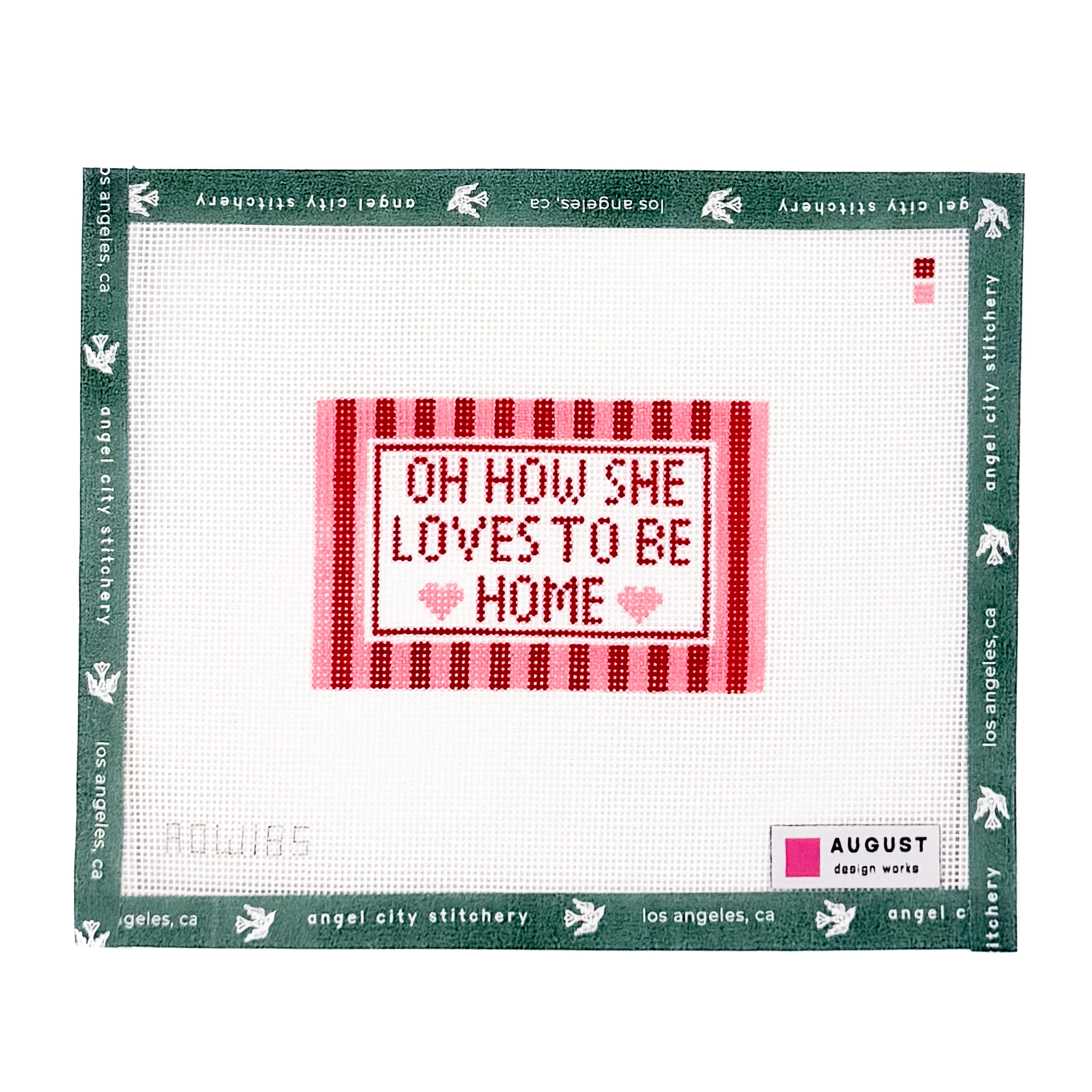 A square counted-stitch canvas featuring the phrase 'OH HOW SHE LOVES TO BE HOME' in red and pink stitched letters on a white grid background, surrounded by a green border printed with 'angel city stitchery' and 'August'.