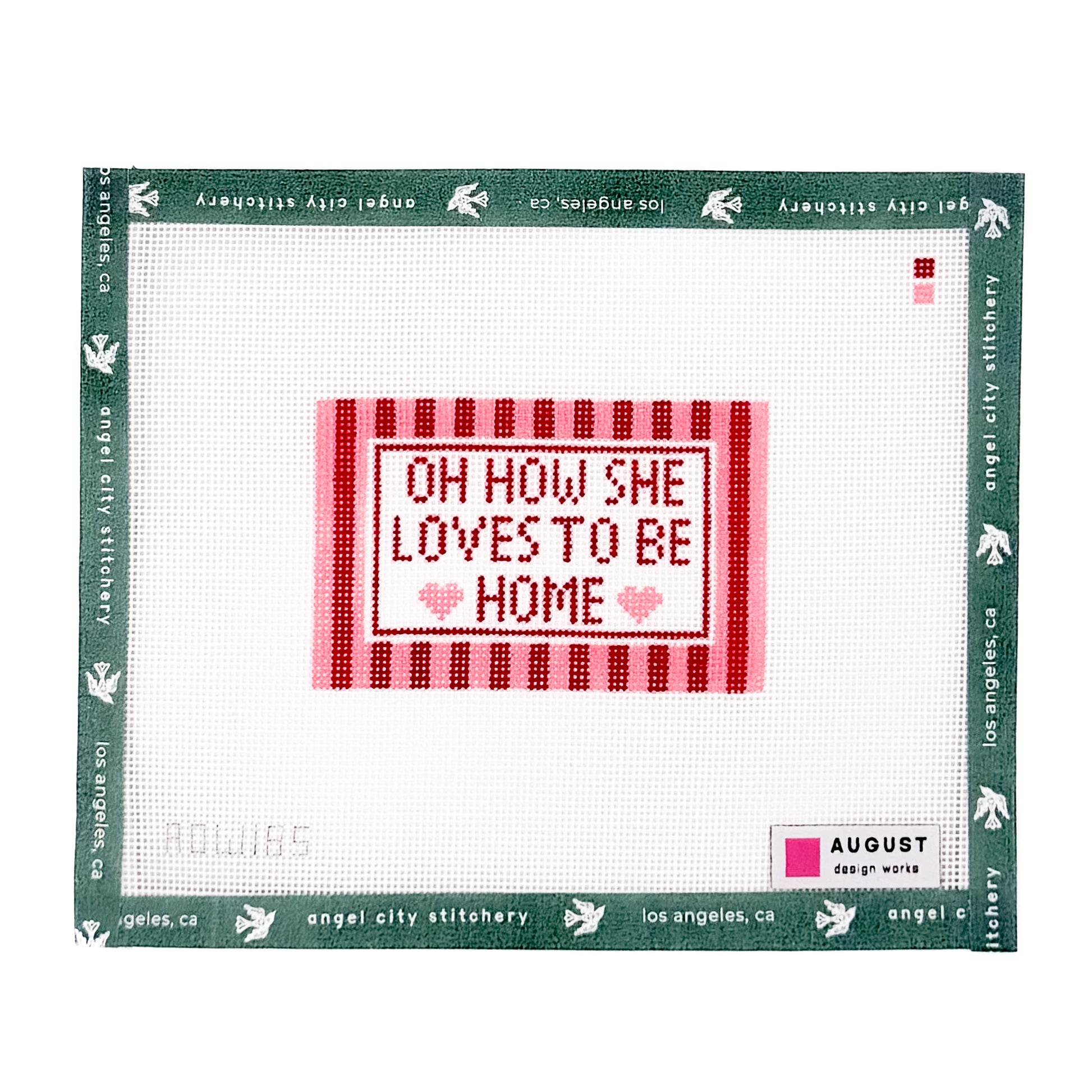 A square counted-stitch canvas featuring the phrase 'OH HOW SHE LOVES TO BE HOME' in red and pink stitched letters on a white grid background, surrounded by a green border printed with 'angel city stitchery' and 'August'.